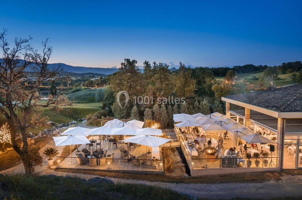 Terrasse éclairée d'un restaurant en soirée, entourée de végétation et offrant une vue sur un paysage vallonné.