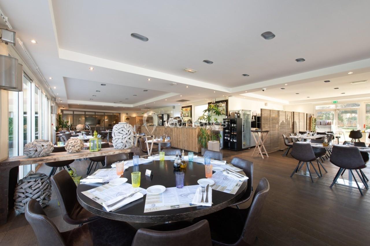 Salle de restaurant lumineuse avec tables dressées, chaises modernes et grandes baies vitrées donnant sur l'extérieur.