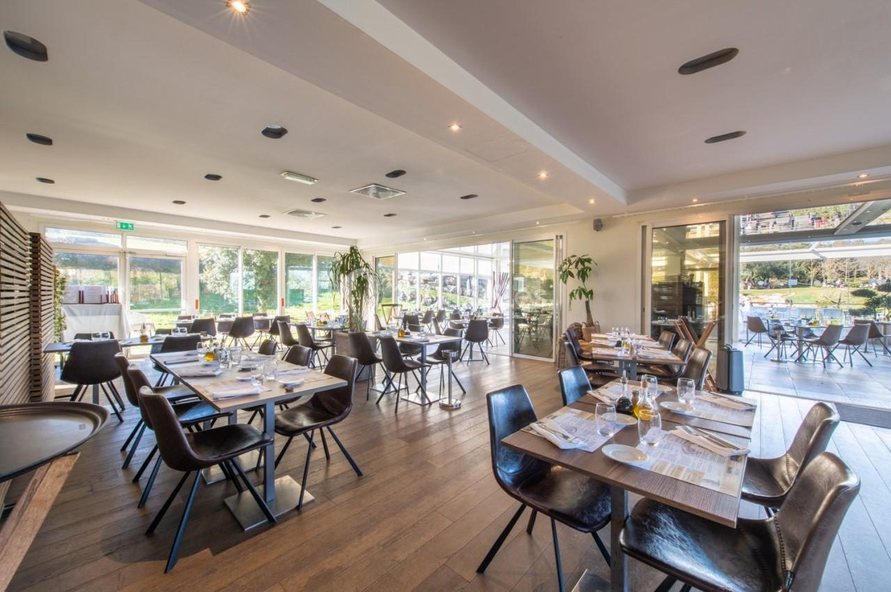 Salle de restaurant lumineuse avec des tables dressées, grandes baies vitrées et vue sur un espace extérieur verdoyant.