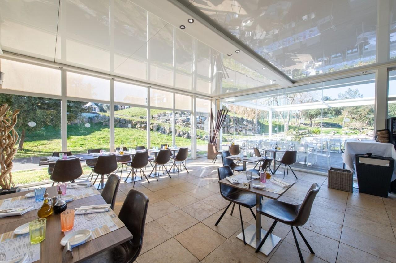 Salle de restaurant lumineuse avec grandes baies vitrées donnant sur un espace extérieur verdoyant.