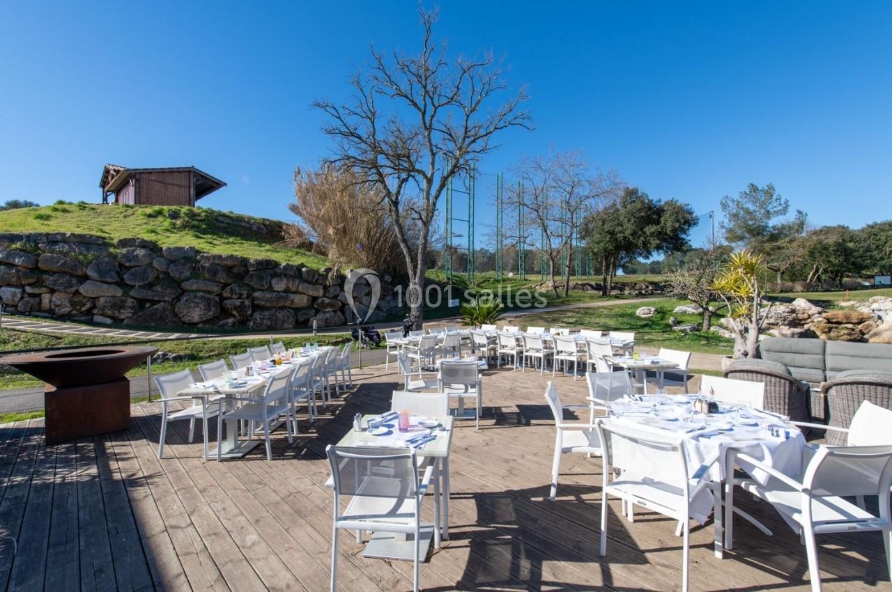 Terrasse en bois aménagée avec des tables et chaises blanches, entourée de verdure et d'arbres sous un ciel dégagé.