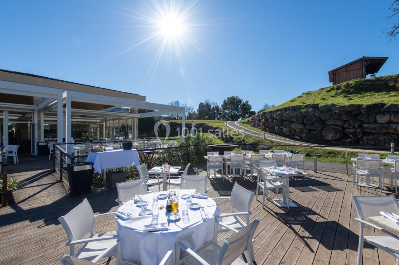 Terrasse ensoleillée d'un restaurant avec tables dressées, vue sur un chemin et une colline verdoyante.