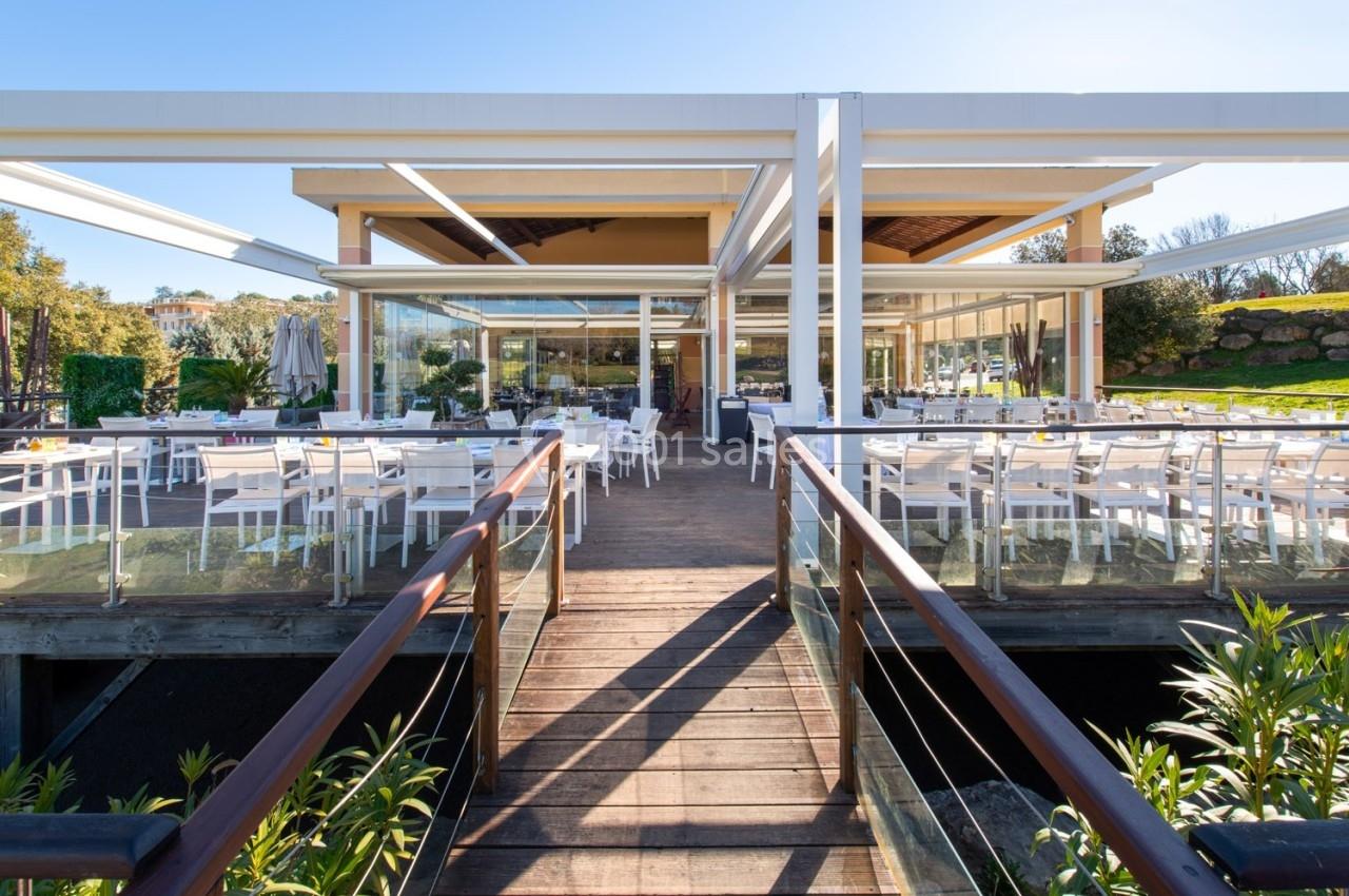 Terrasse d'un restaurant moderne avec tables et chaises blanches, entourée de verdure et éclairée par la lumière naturelle.