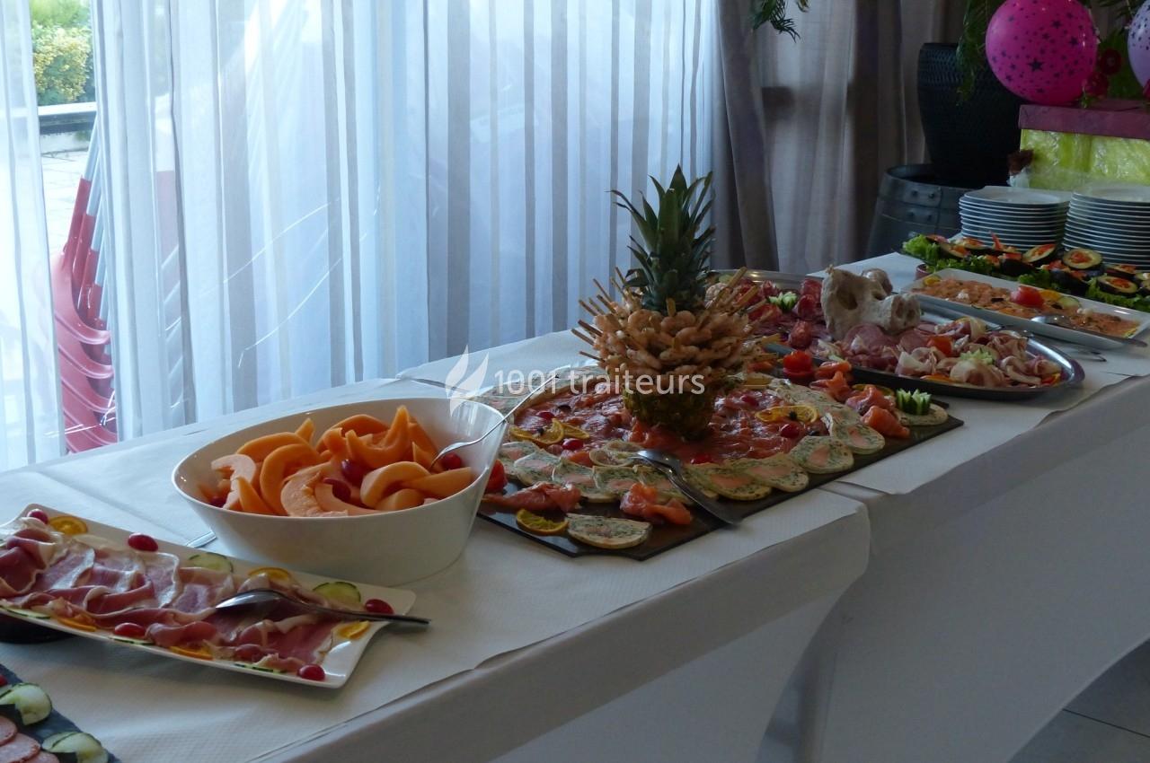 Buffet avec des plats variés comprenant fruits, charcuterie et légumes, disposés sur une table blanche près d'une fenêtre.