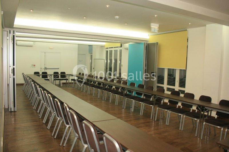 Salle de réunion lumineuse avec de longues tables en U, des chaises alignées et un sol en parquet.