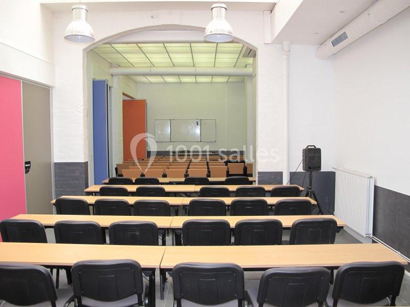Salle de classe vide avec des rangées de tables et chaises, tableau blanc au fond et éclairage au plafond.