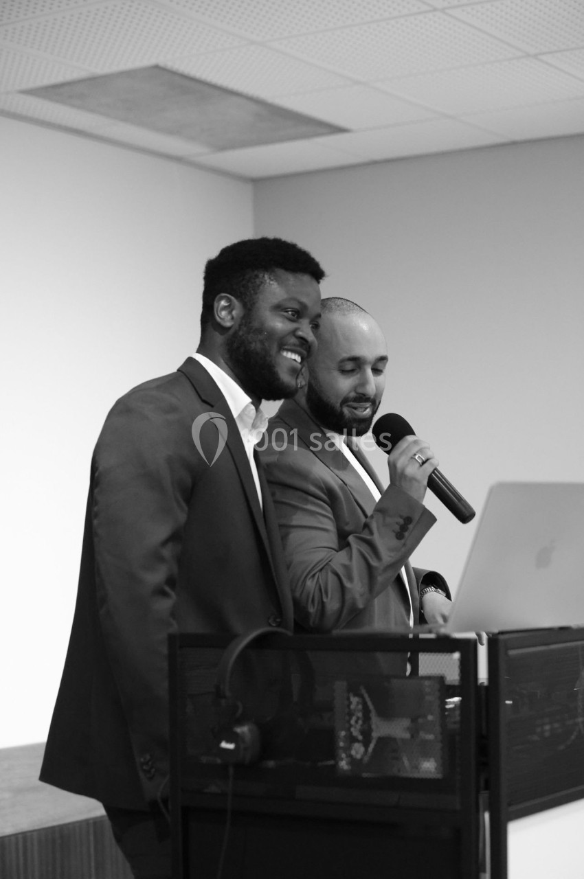 Deux hommes en costume parlent et sourient devant un ordinateur portable, l'un tenant un micro.