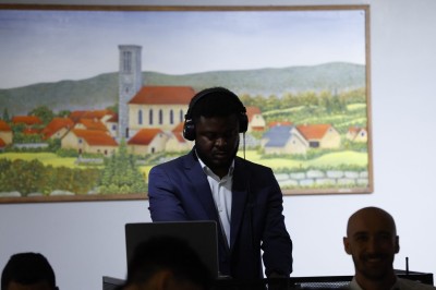 Un DJ en costume mixe de la musique devant une peinture représentant un village avec des collines en arrière-plan.