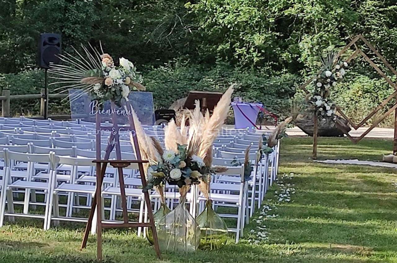 Chaises blanches alignées en extérieur pour une cérémonie, décorations florales et panneau ’Bienvenue’ au premier plan.
