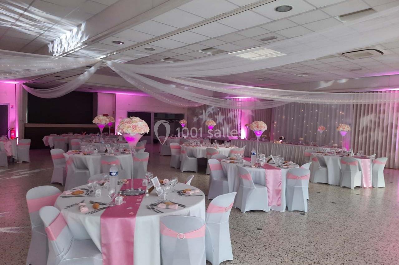 Salle de réception décorée avec des nappes blanches et roses, des centres de table fleuris et un éclairage rose.