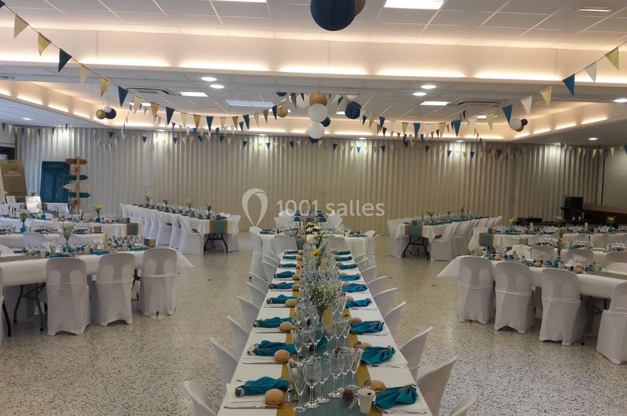 Salle de réception décorée avec des tables alignées, nappes blanches, serviettes turquoise et guirlandes suspendues.
