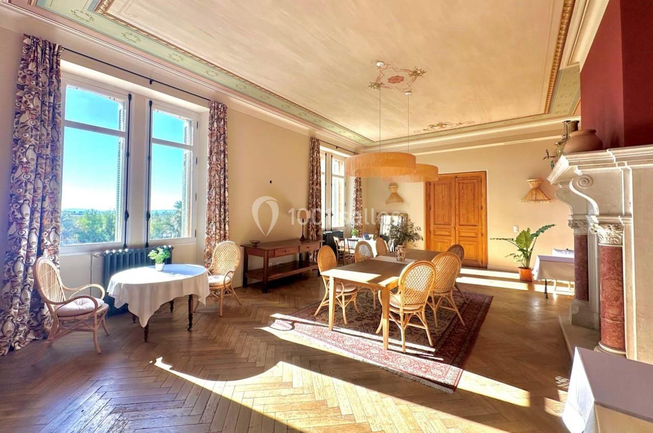 Salle lumineuse avec parquet en bois, grandes fenêtres, tables et chaises en rotin, et cheminée décorative.