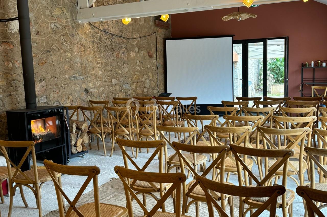 Salle de réunion avec des chaises en bois disposées face à un écran de projection, dans un cadre rustique avec mur en pierre.