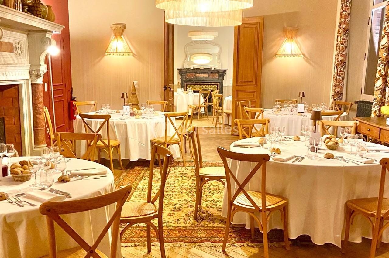 Salle de réception élégante avec tables rondes dressées, chaises en bois et éclairage chaleureux.
