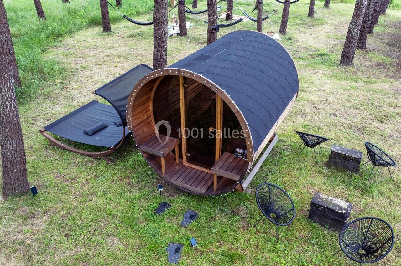 Sauna en bois en forme de tonneau installé dans une clairière boisée, entouré de chaises et de hamacs.