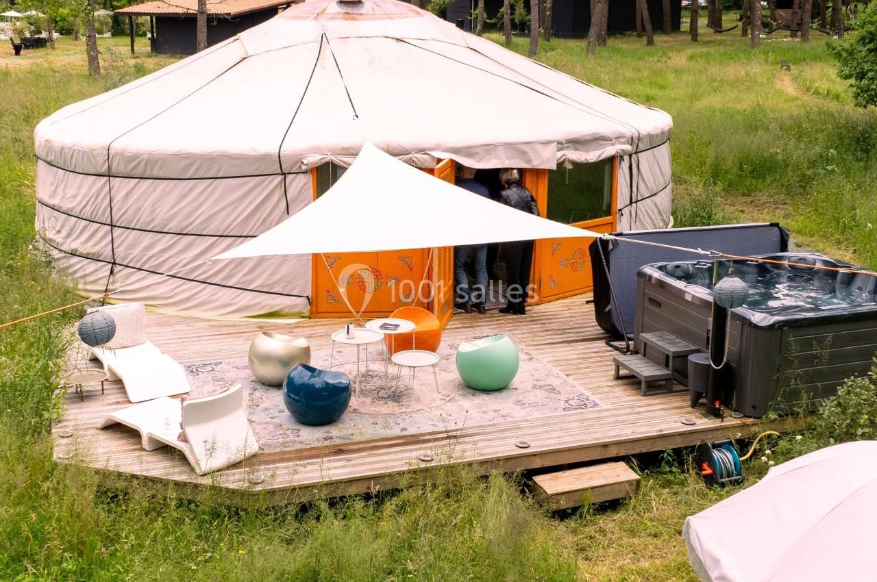 Yourte avec terrasse en bois aménagée, mobilier extérieur, parasol et jacuzzi, entourée de verdure et d'arbres.