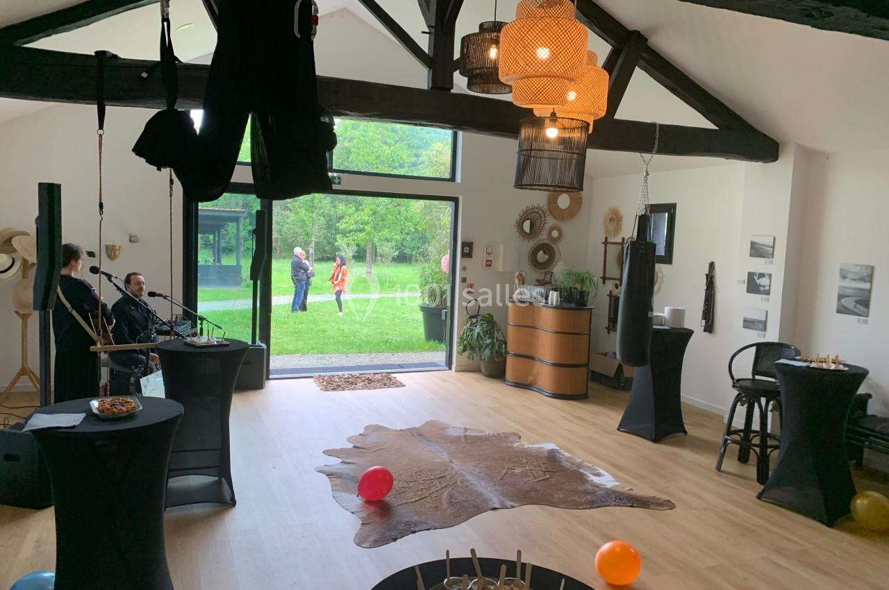 Salle lumineuse avec poutres apparentes, décorée pour un événement, vue sur un jardin à travers de grandes baies vitrées.