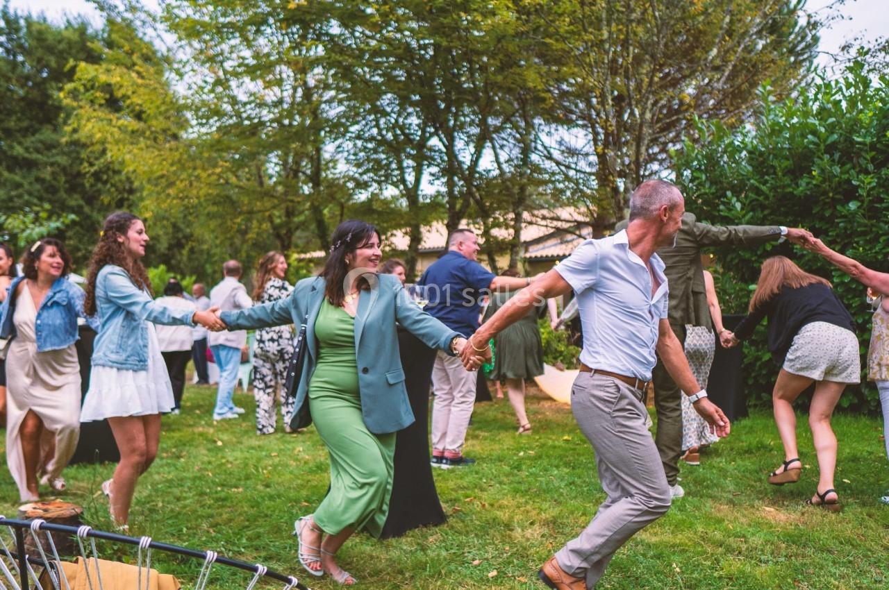 Des personnes dansent en chaîne en extérieur sur une pelouse, entourées d'arbres et de tables de réception.