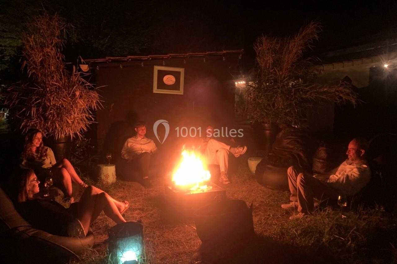 Personnes assises autour d'un feu de camp dans un jardin, éclairées par les flammes et des lumières tamisées.