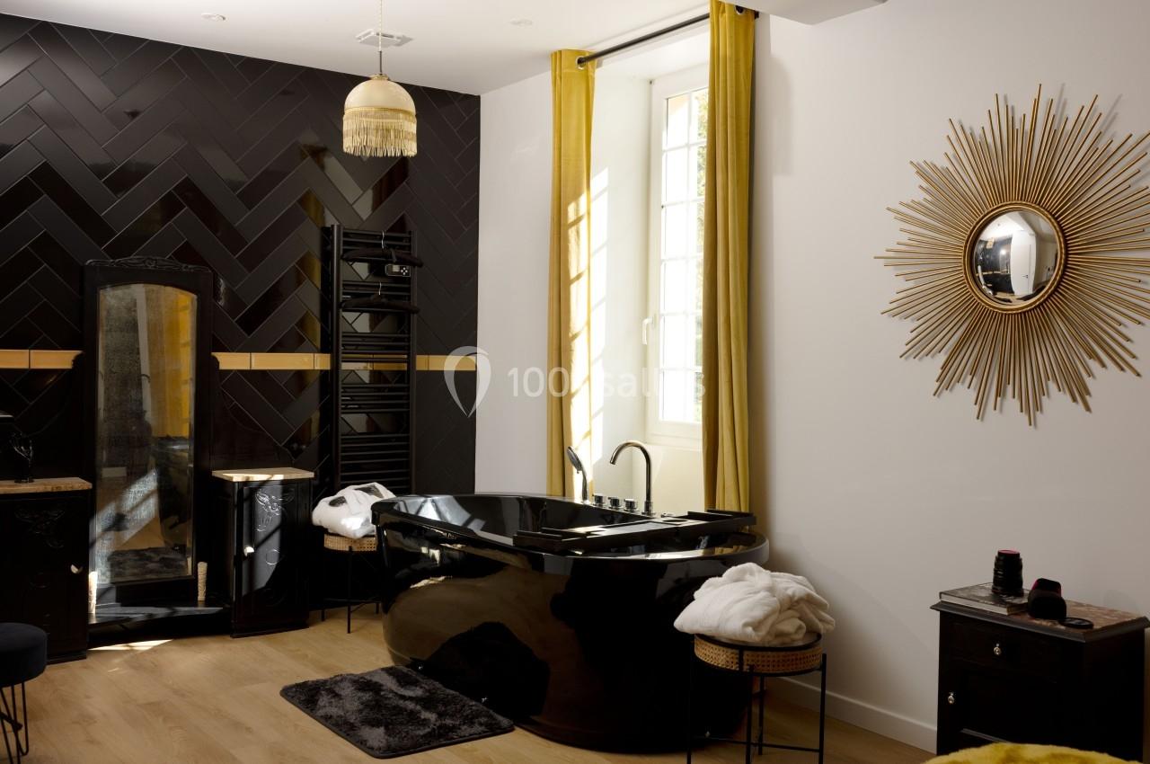 Salle de bain moderne avec baignoire noire, miroir doré en forme de soleil et mur en carrelage noir en chevrons.