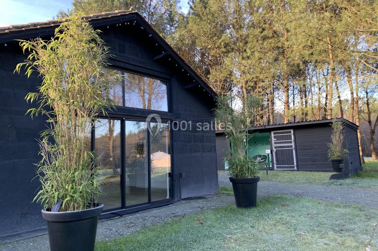 Maison en bois noir avec grandes baies vitrées, entourée de bambous en pots et d'arbres dans un cadre naturel.