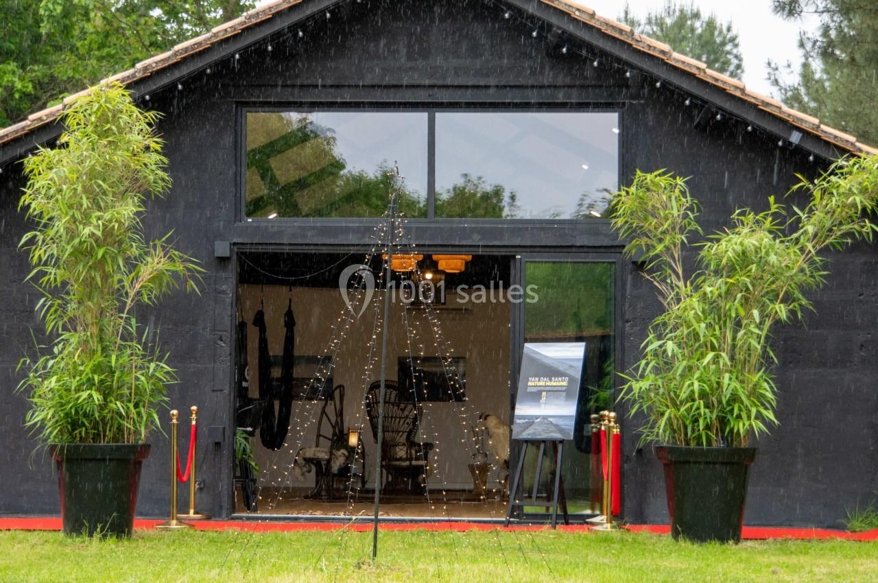 Façade d'un bâtiment noir avec de grandes baies vitrées, encadré par deux plantes en pot et éclairé à l'intérieur.