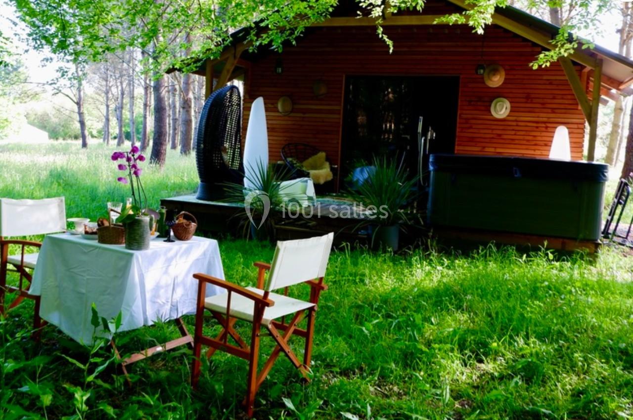 Cabane en bois avec terrasse, entourée de verdure, table dressée et fauteuils pliants sur une pelouse.