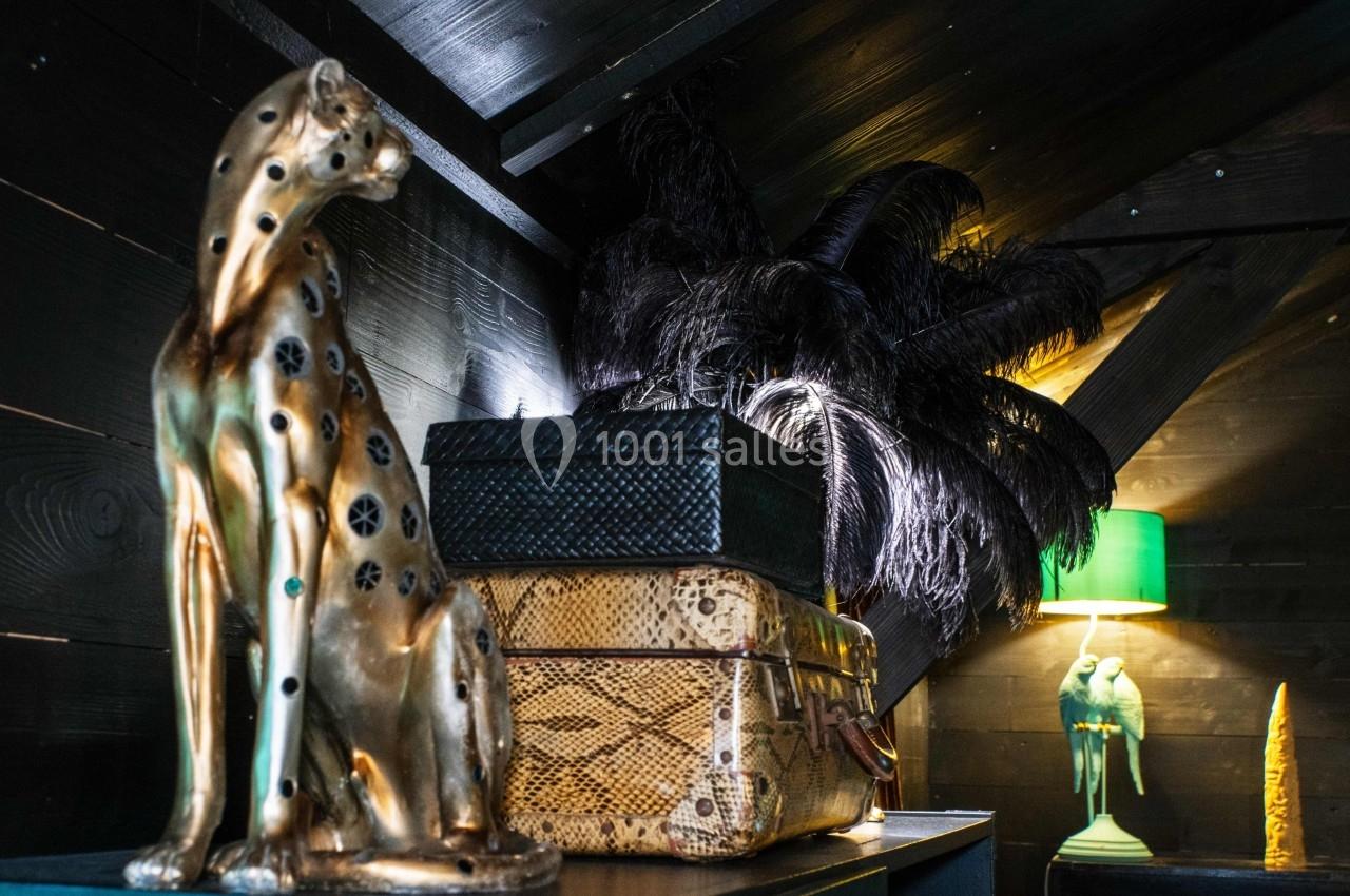 Statue dorée de guépard, boîtes décoratives et lampe verte dans un intérieur sombre avec plumes noires en arrière-plan.