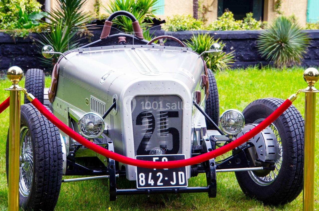 Voiture de course vintage argentée exposée sur une pelouse, entourée de cordons rouges et de poteaux dorés.