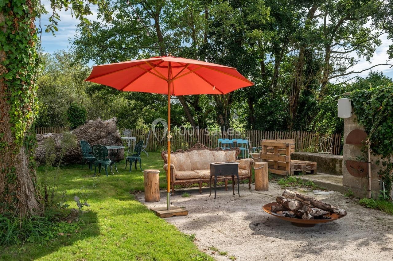 Espace extérieur avec canapé, parasol rouge, brasero et mobilier de jardin entouré de verdure et d'arbres.