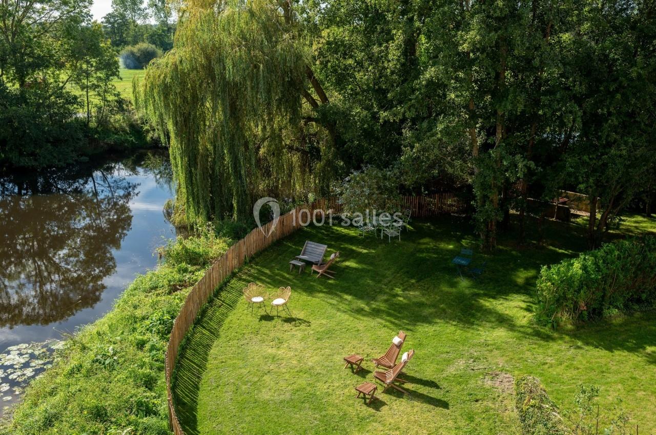 Jardin verdoyant avec des chaises longues, une table et des arbres, bordé par une rivière calme et une clôture en bois.