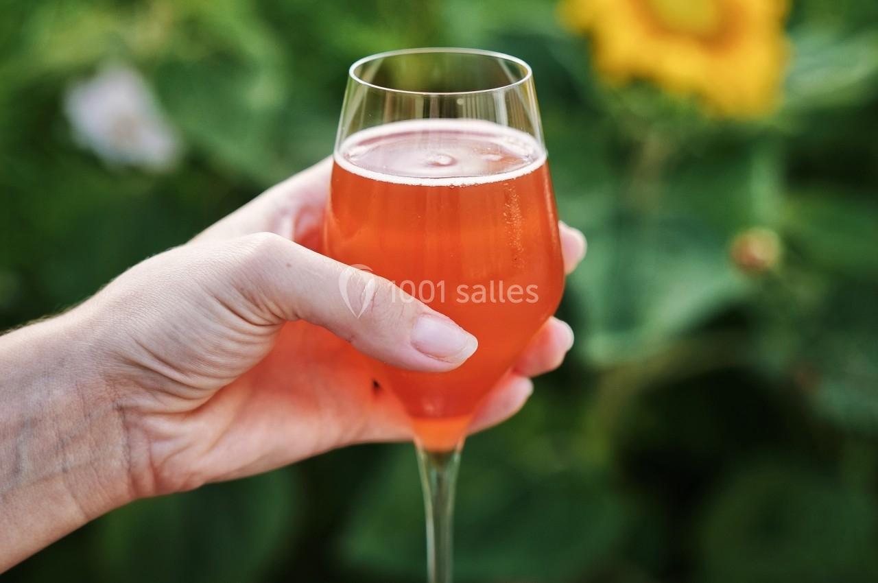 Un verre de boisson orange tenu par une main, avec des tournesols et un feuillage en arrière-plan.