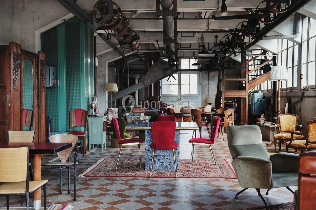 Salle industrielle aménagée avec des meubles éclectiques, incluant des chaises colorées et des tables sur un sol en carreaux.