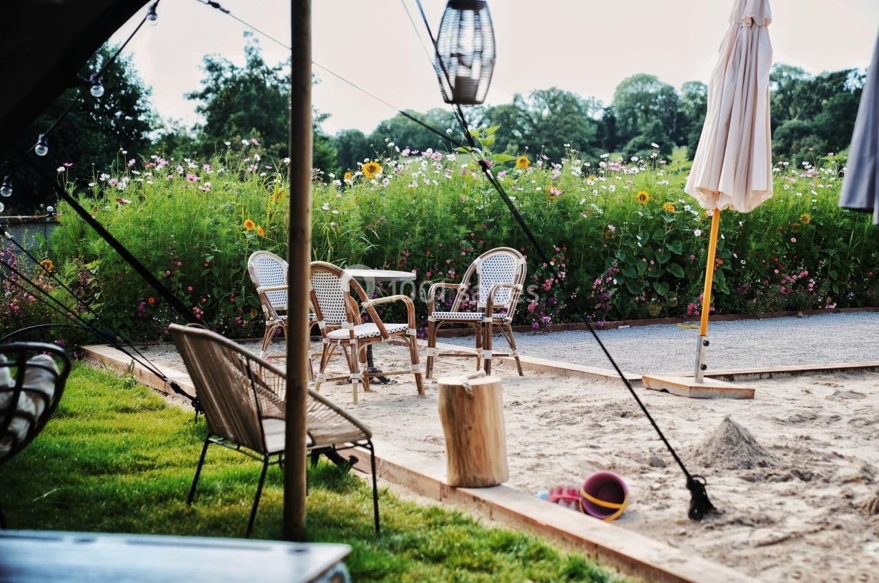 Espace extérieur avec chaises en rotin, table, parasol et jouets dans un bac à sable, entouré de fleurs et verdure.