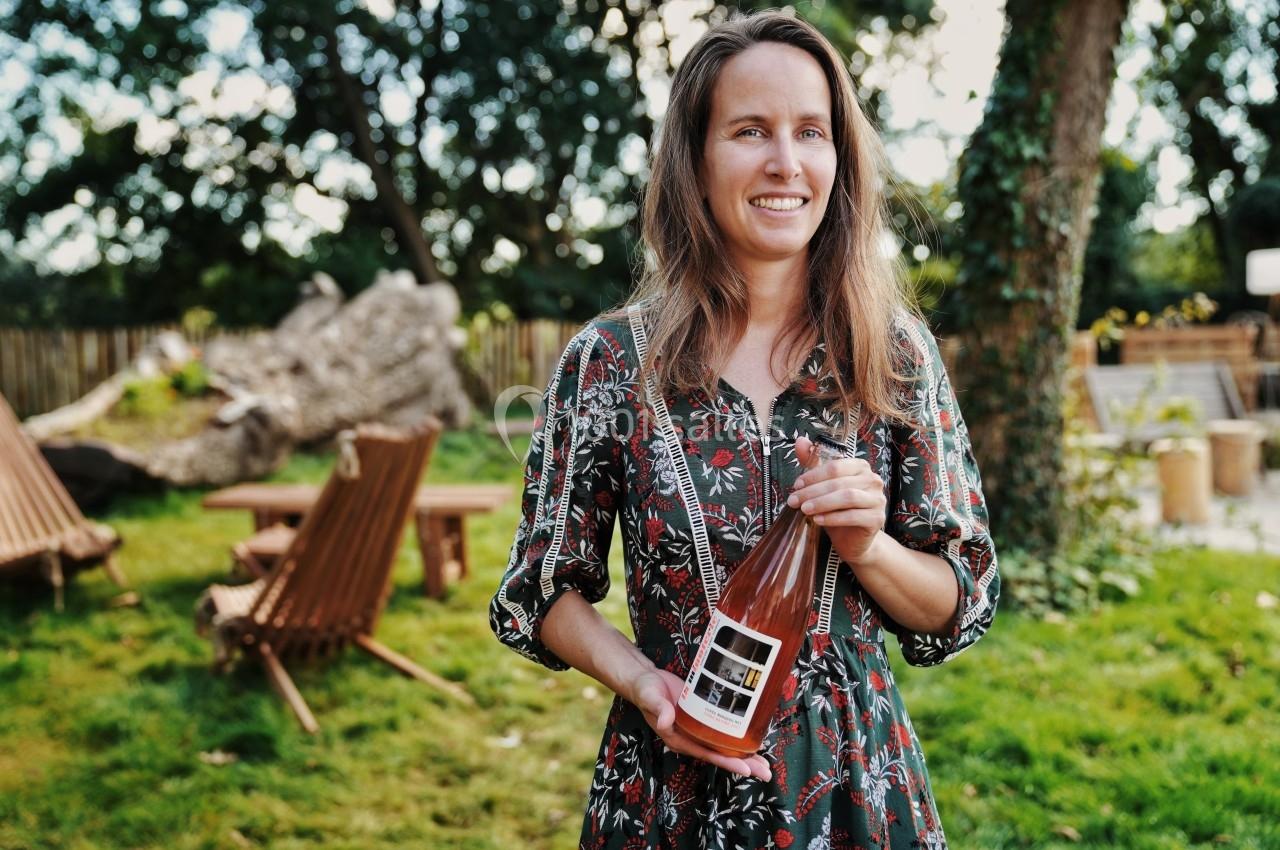 Une femme souriante tient une bouteille de boisson rosée dans un jardin avec des chaises en bois en arrière-plan.