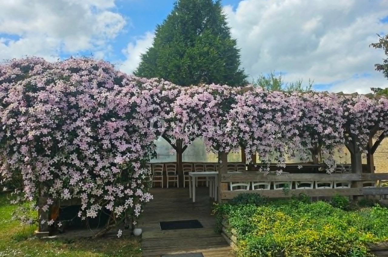 Pergola en bois couverte de fleurs roses, entourée de verdure, avec des tables et chaises disposées dessous.