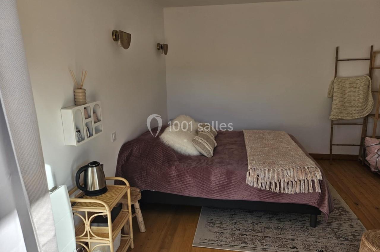 Chambre lumineuse avec lit double, plaid beige, coussins moelleux, étagère murale et petit coin avec bouilloire.