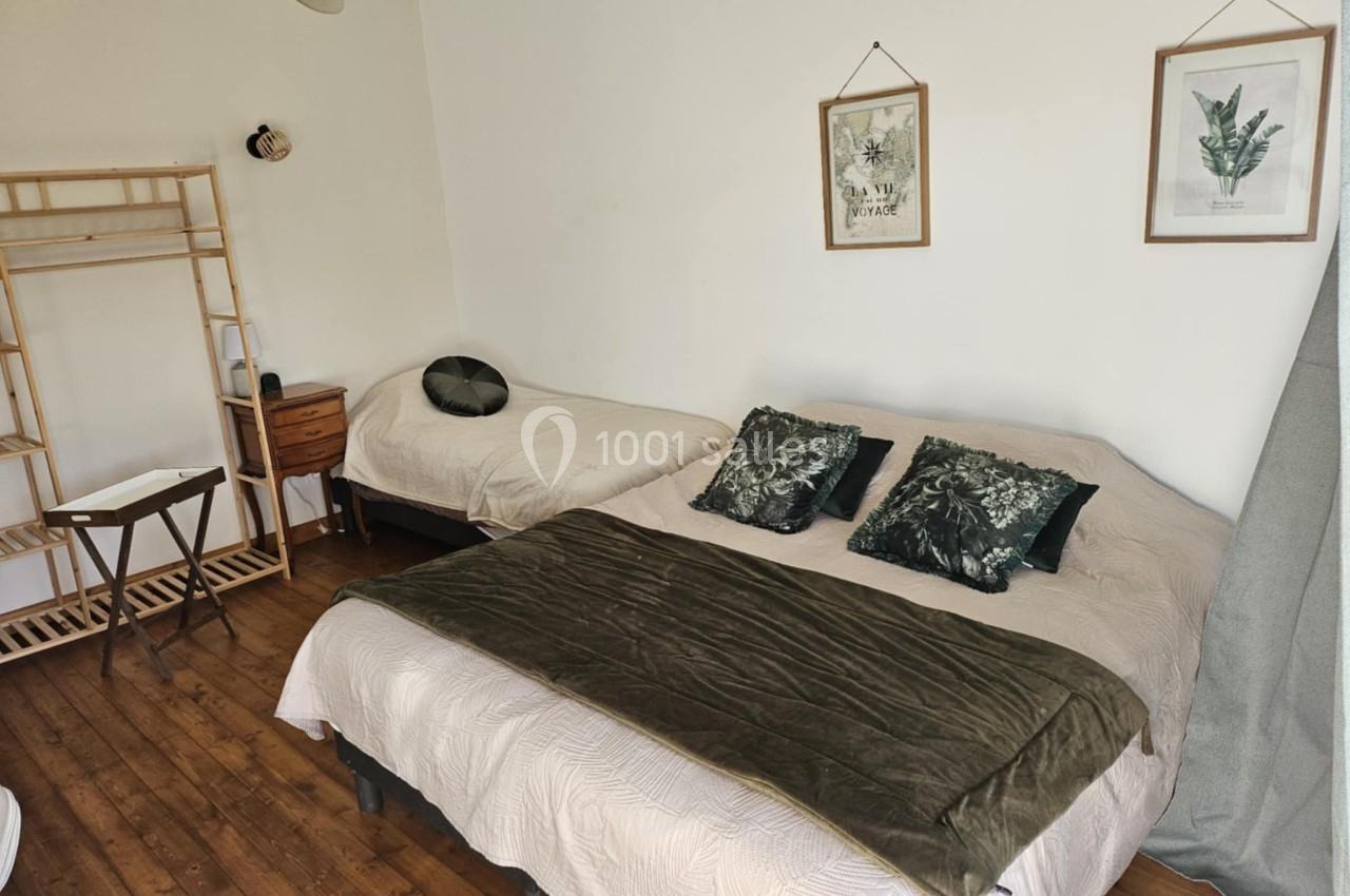 Chambre avec un lit double, un lit simple, des coussins verts, un portant en bois et des cadres végétaux au mur.