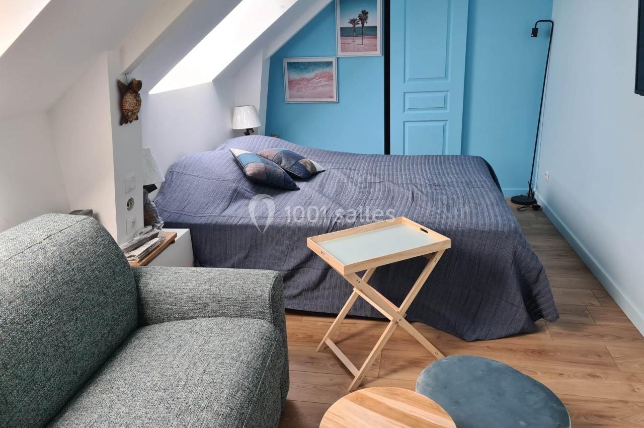Chambre mansardée avec lit double, canapé, table pliante, pouf et décoration murale sur fond bleu clair.