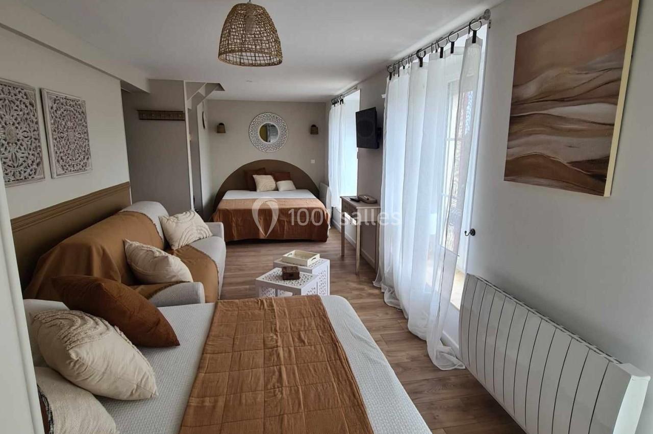 Chambre lumineuse avec deux lits, canapé, parquet en bois, décoration neutre et grande fenêtre avec rideaux blancs.