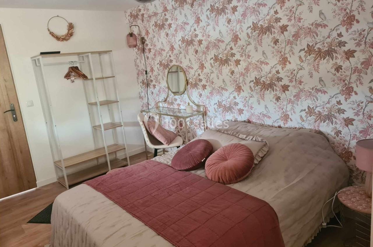 Chambre avec lit double, coussins roses, papier peint floral, coiffeuse et portant en bois dans un décor chaleureux.