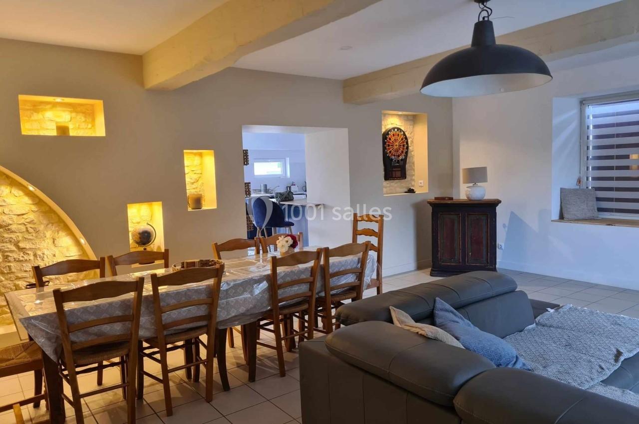 Salle à manger lumineuse avec table en bois, chaises assorties, mur en pierre éclairé et salon attenant.