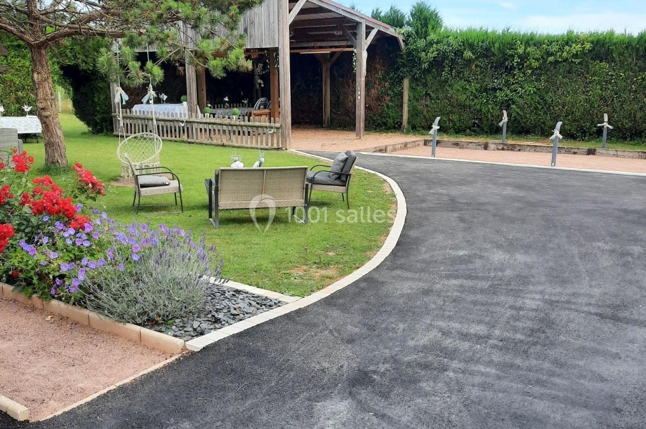 Allée asphaltée menant à un espace extérieur aménagé avec pelouse, fleurs, bancs et abri en bois.