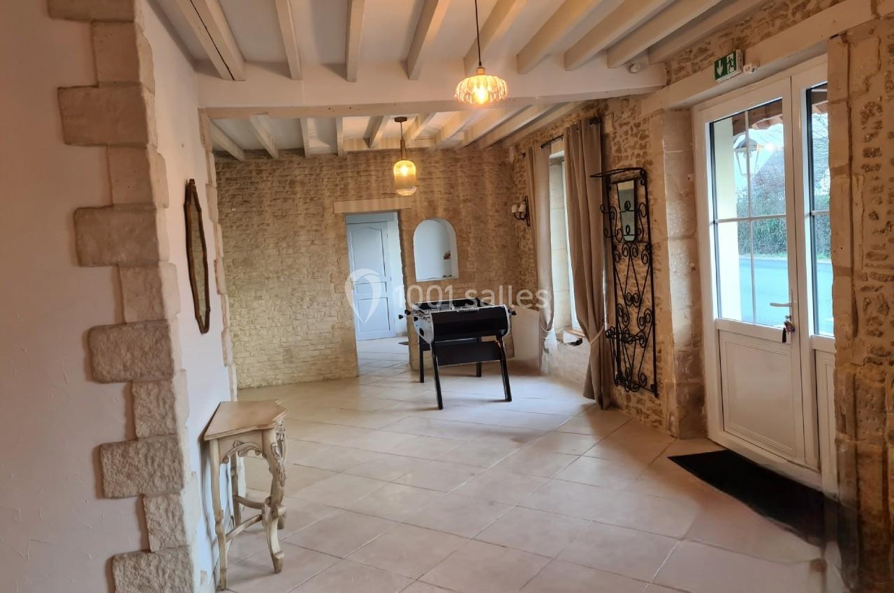 Salle lumineuse avec murs en pierre, plafond à poutres apparentes, baby-foot et porte-fenêtre donnant sur l'extérieur.