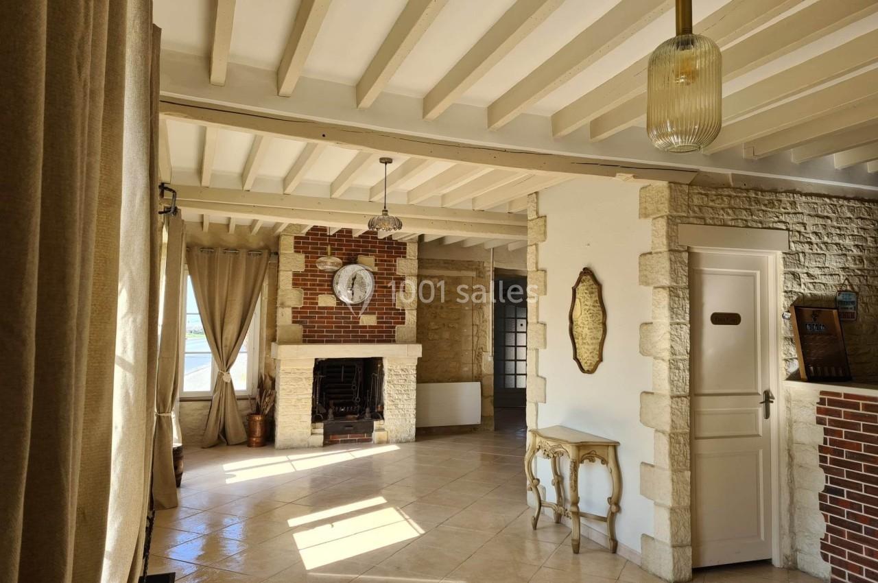 Intérieur lumineux avec cheminée en briques, murs en pierre, poutres apparentes et grandes fenêtres avec rideaux beiges.