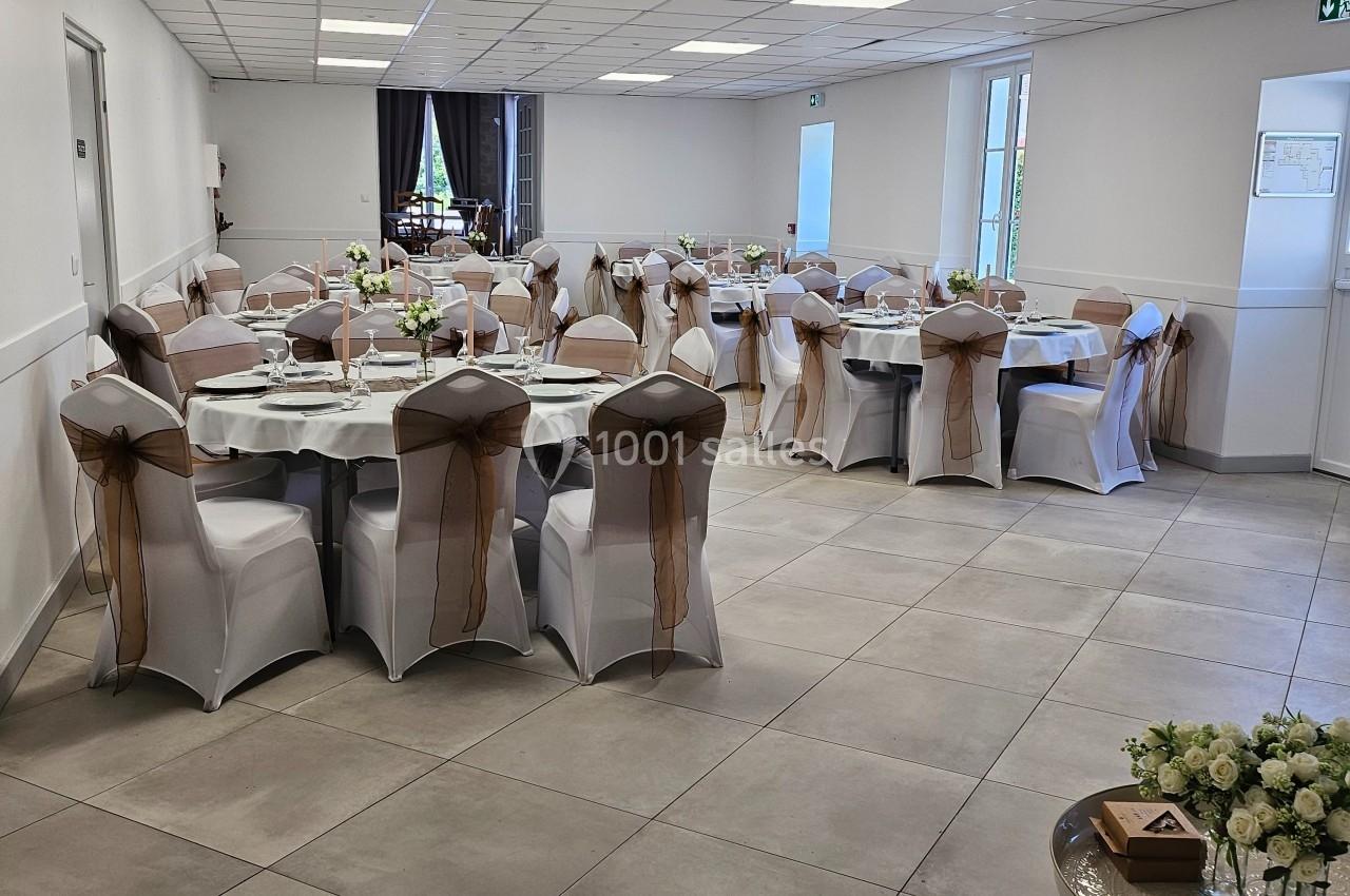 Salle de réception lumineuse avec tables rondes dressées, chaises décorées de nœuds marron et bouquets de fleurs.