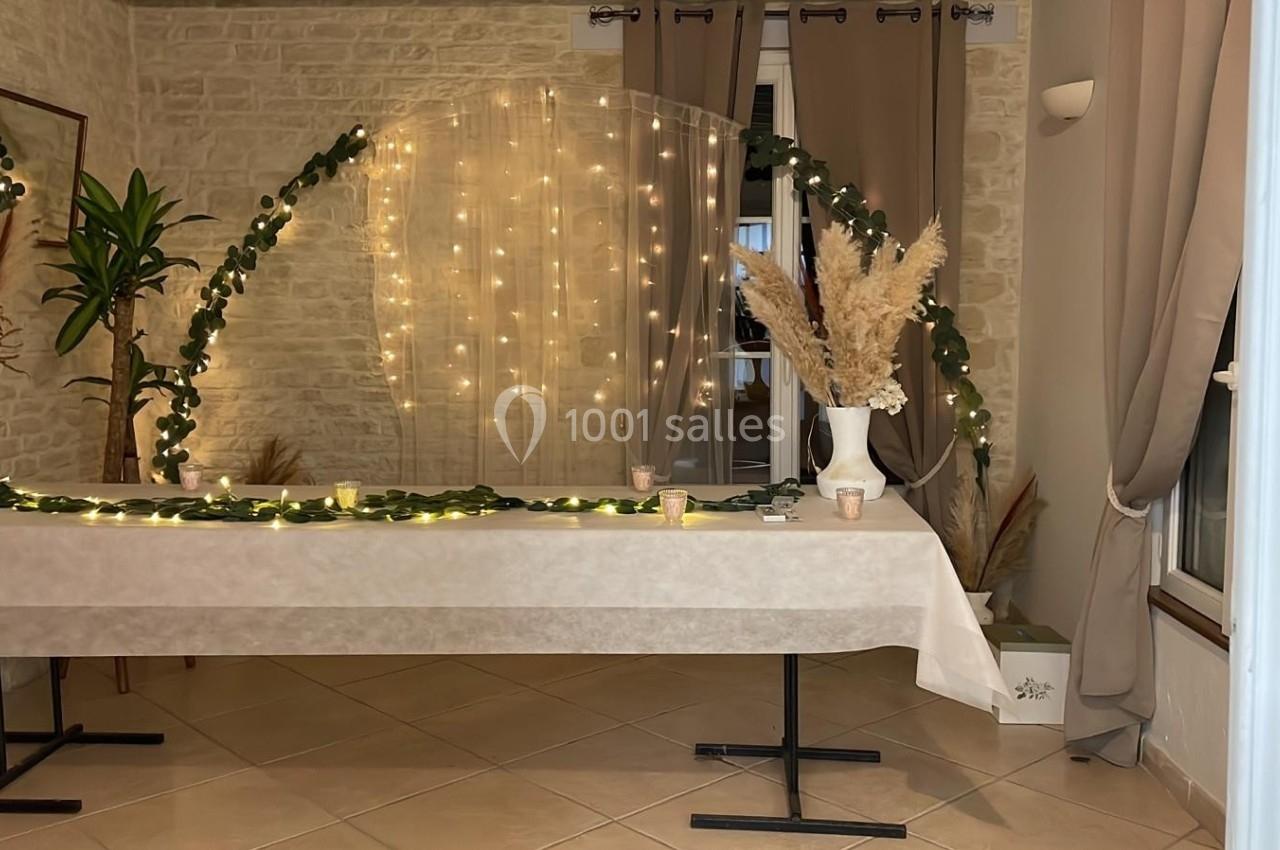 Table décorée avec une nappe blanche, guirlandes lumineuses, feuillage, vases avec pampas, dans une pièce aux murs en pierre.
