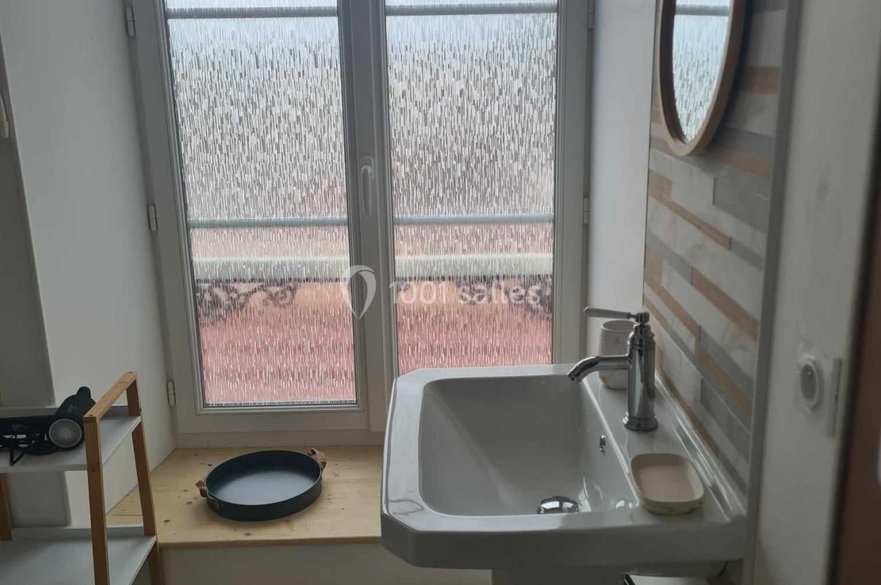 Lavabo blanc avec robinet, miroir rond au mur, étagère en bois et fenêtre avec vitrage dépoli en arrière-plan.
