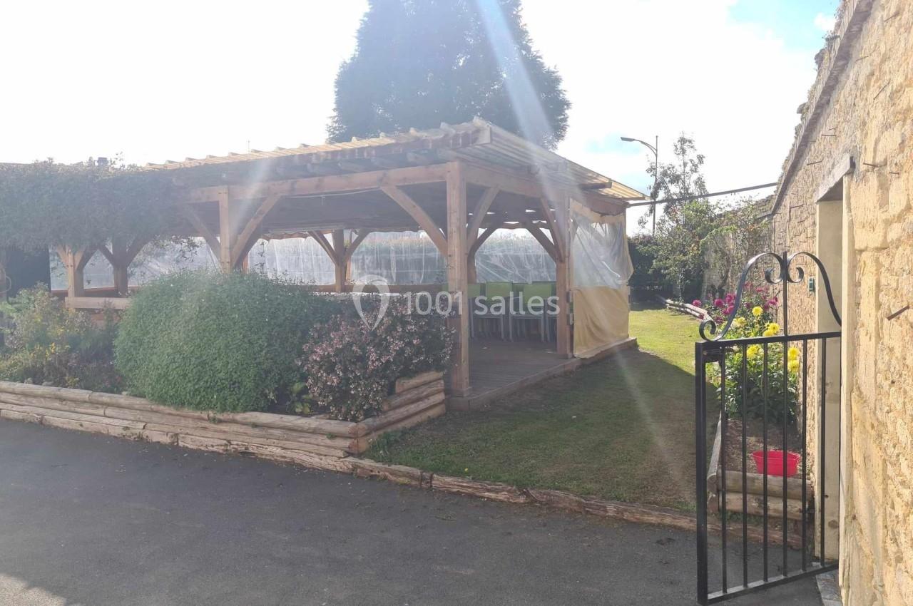 Abri en bois avec table et chaises, entouré de plantes, situé dans une cour ensoleillée près d'un mur en pierre.