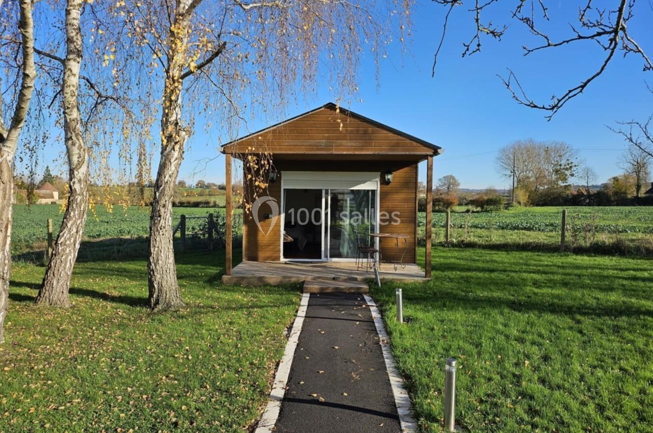 Petit chalet en bois avec une large baie vitrée, entouré d'arbres et situé dans un paysage champêtre ensoleillé.